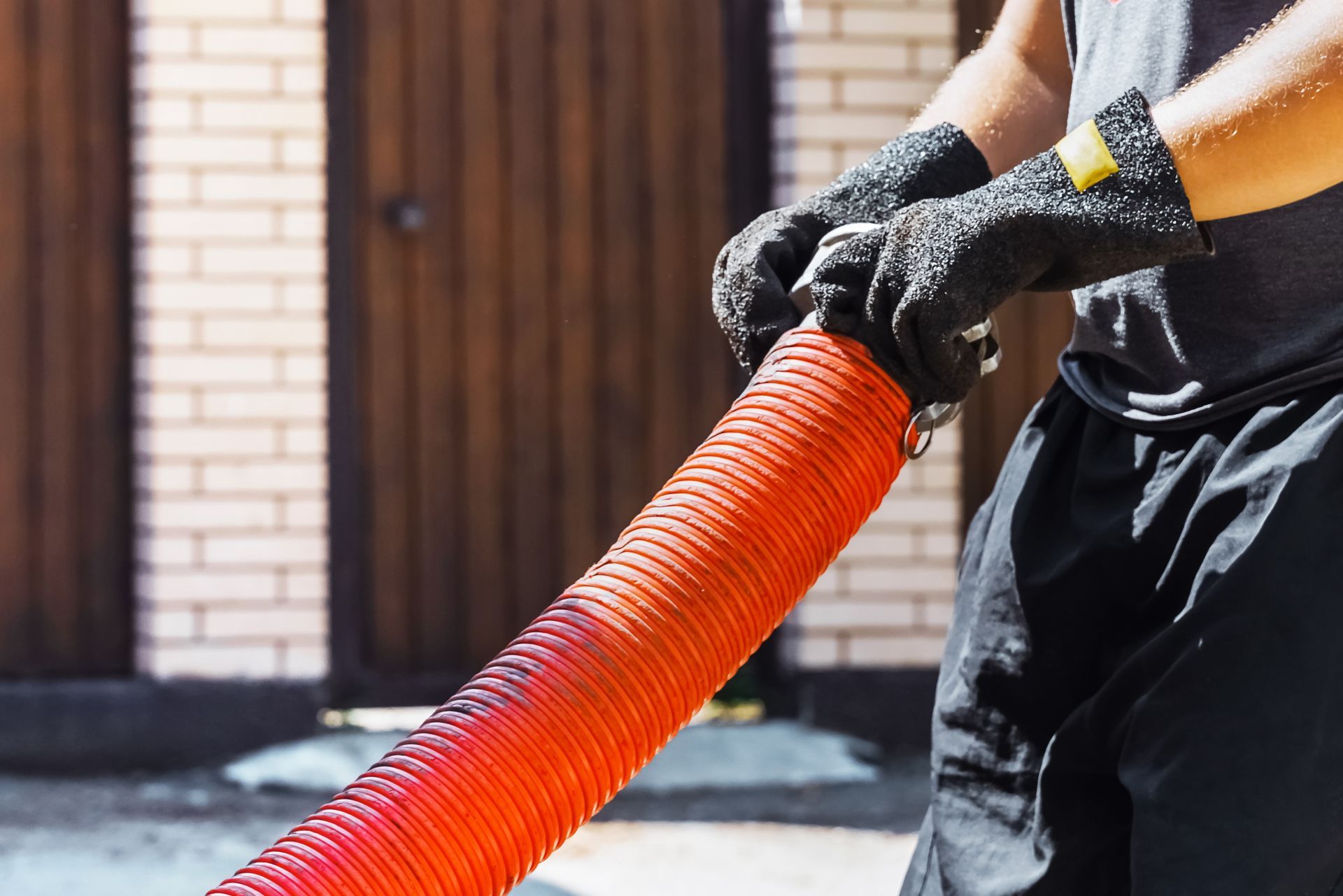 Gloved person holding an orange corrugated hose outdoors near a building