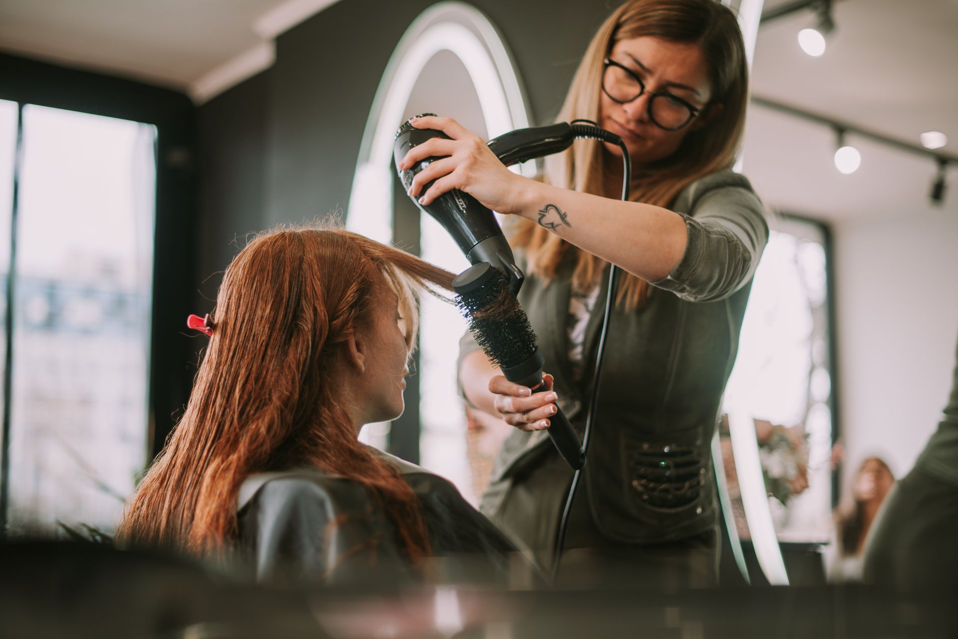 A hairstylist uses a round brush and hairdryer to style a client's textured, copper-colored hair in a salon.