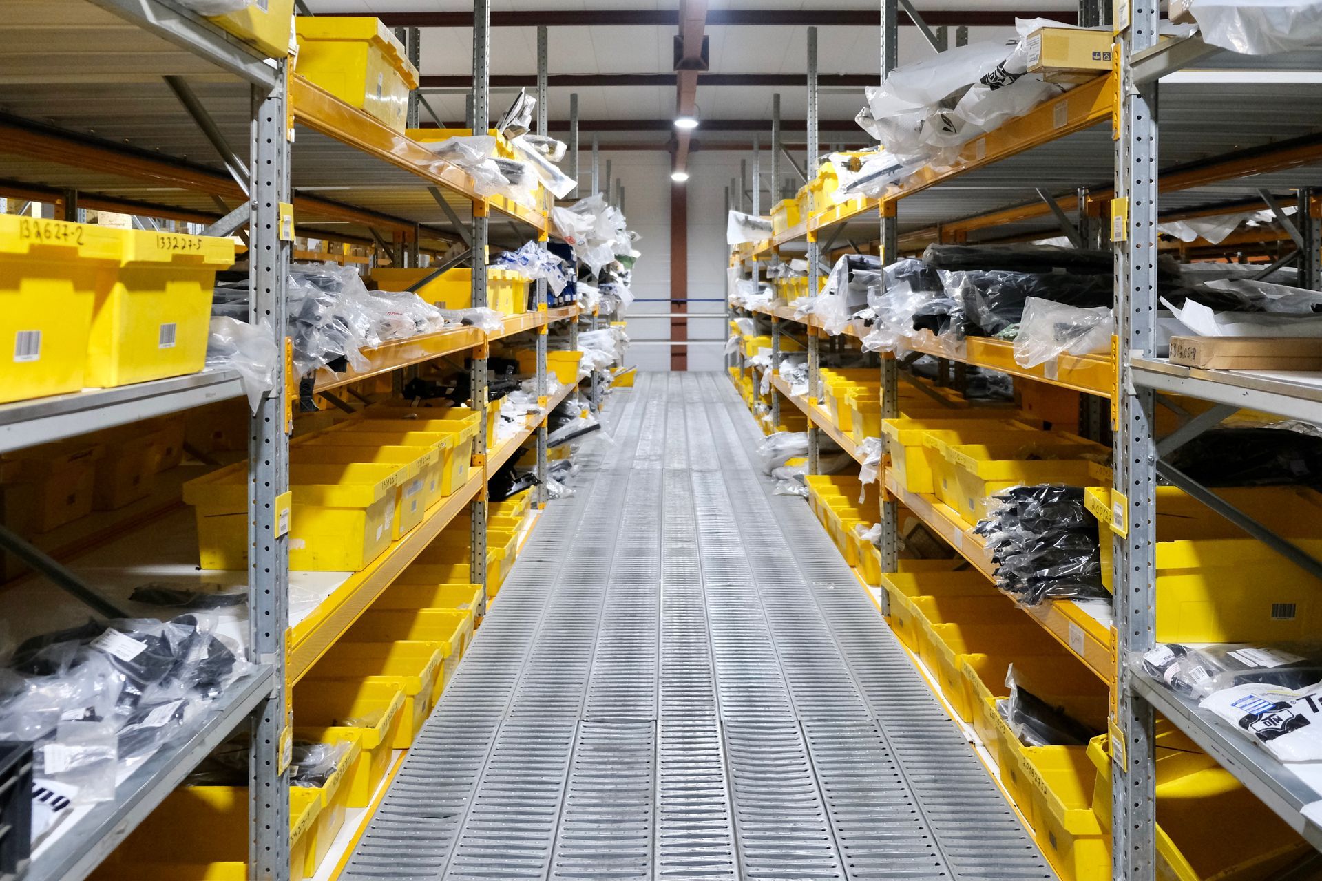 Aisle of warehouse shelves with yellow bins and stacked parts on both sides of a metal walkway.