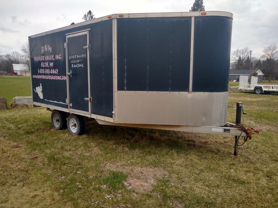 Black enclosed trailer with ramp and side door parked on grass under cloudy sky.
