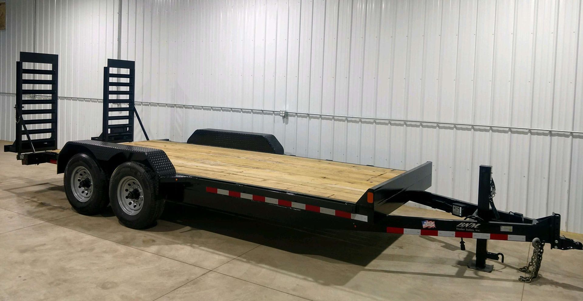 Black dual-axle utility trailer with wooden deck, rail ramps, and hitch in an indoor warehouse.