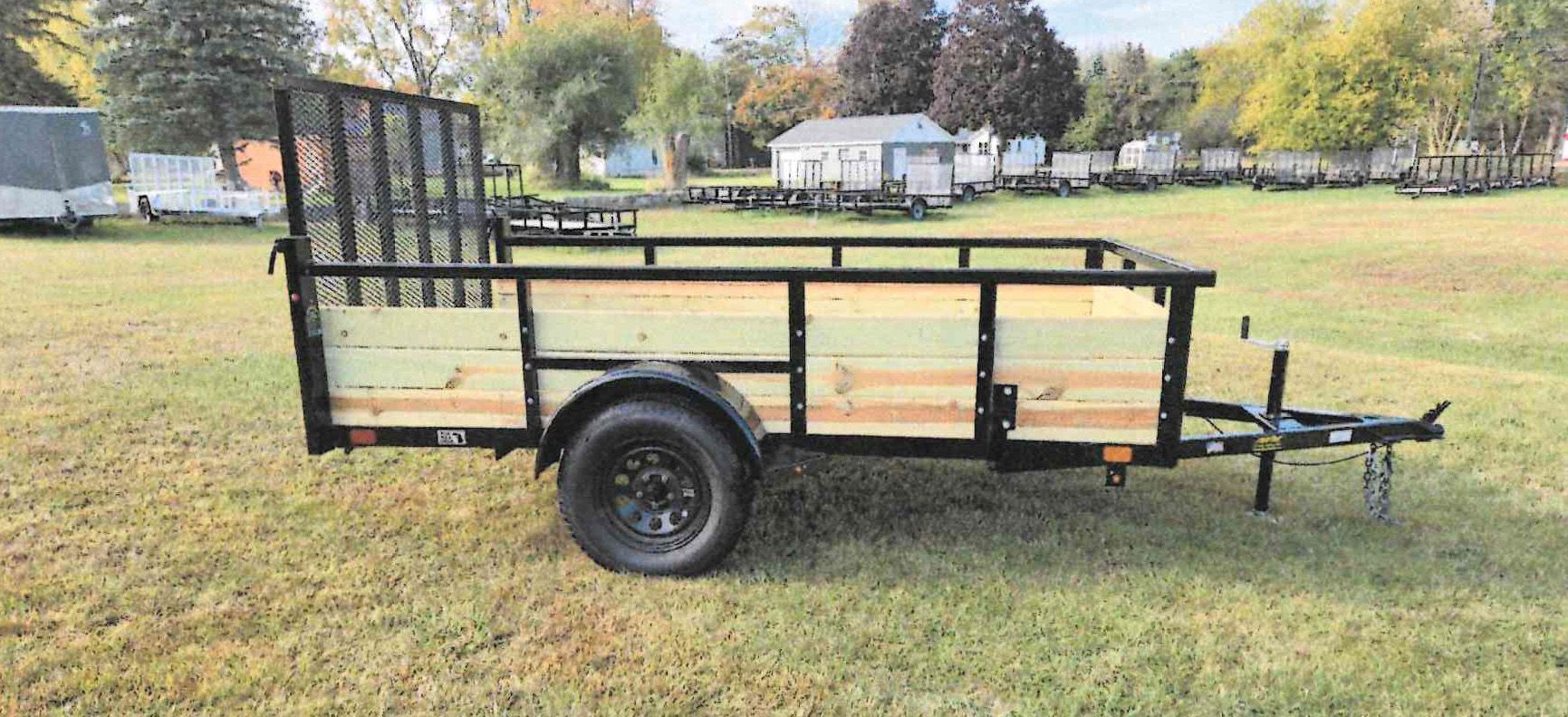 Black utility trailer with side rails and a hitch, parked on grass in a park.