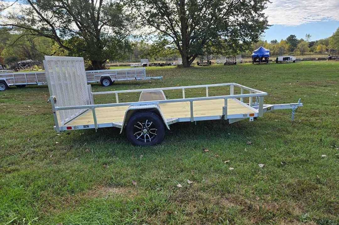 Flatbed utility trailer with mesh sides on a grassy field near trees and parked vehicles.