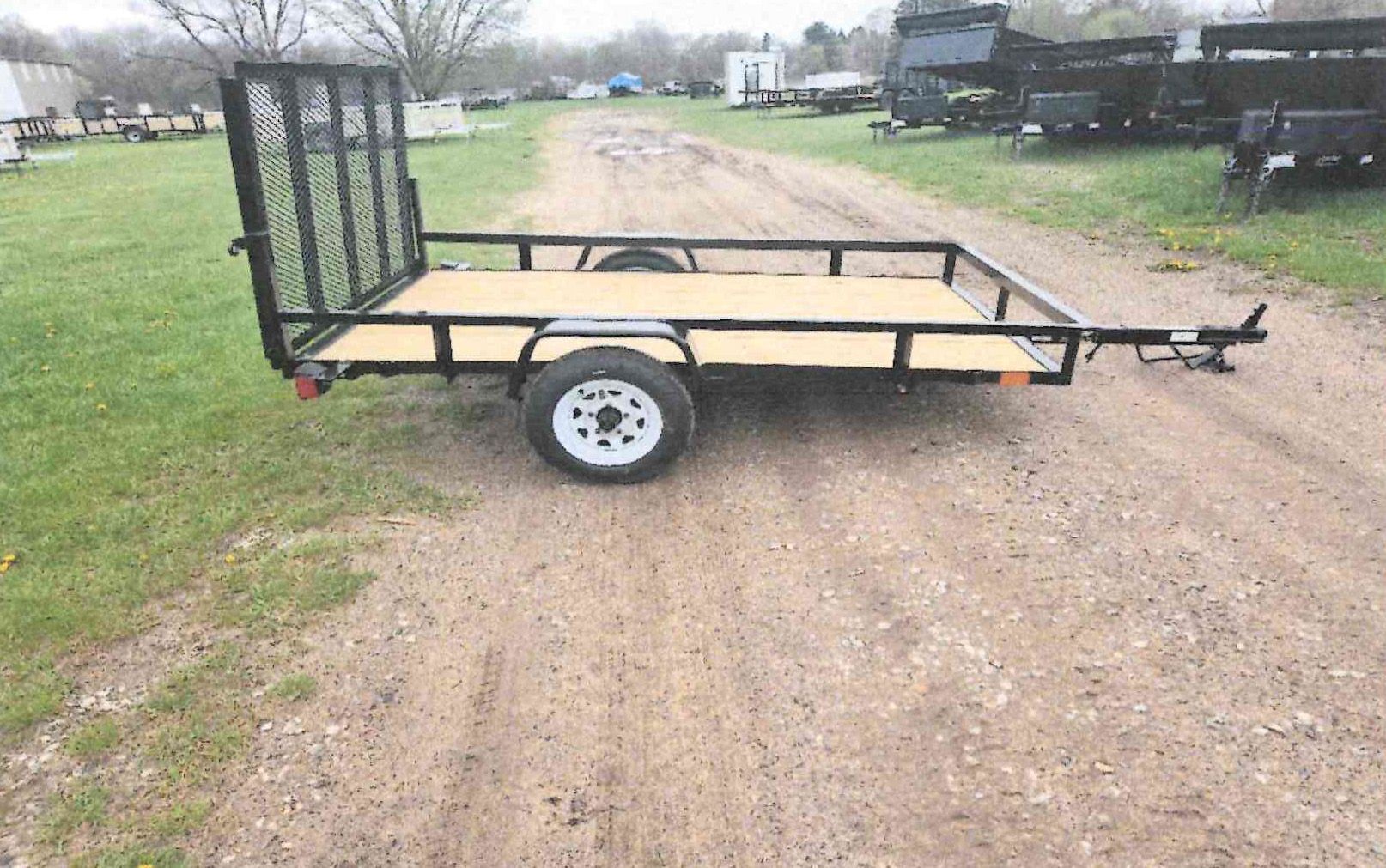 Black utility trailer on a gravel driveway beside grass, with a folded mesh ramp gate.