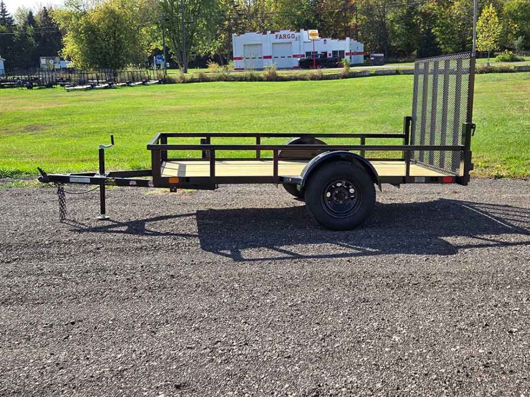 Black utility trailer parked on gravel beside a grassy field and trees under a clear sky