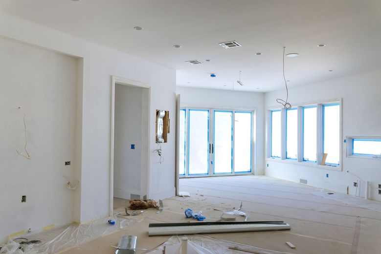 A bright, unfinished room with white walls, recessed ceiling lights, large windows, and floor protection paper.