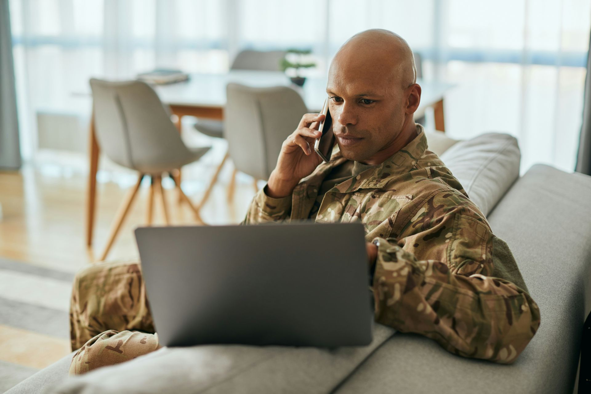A soldier is sitting on a couch using a laptop and talking on a cell phone.