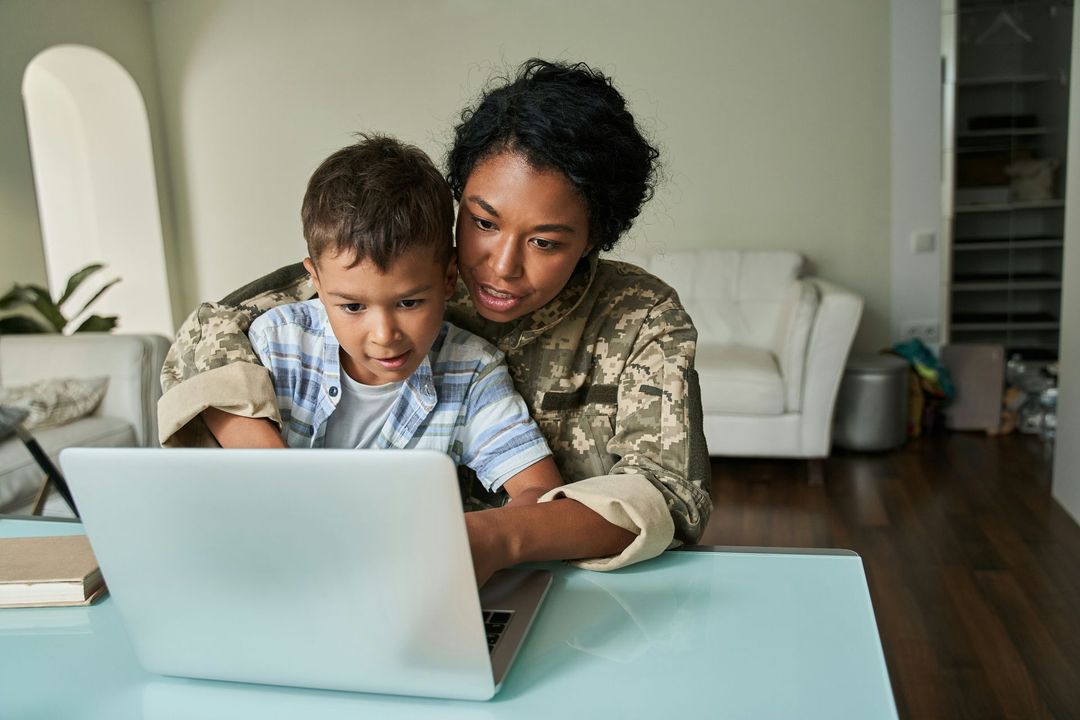 A woman in a military uniform is sitting at a table with a child looking at a laptop.