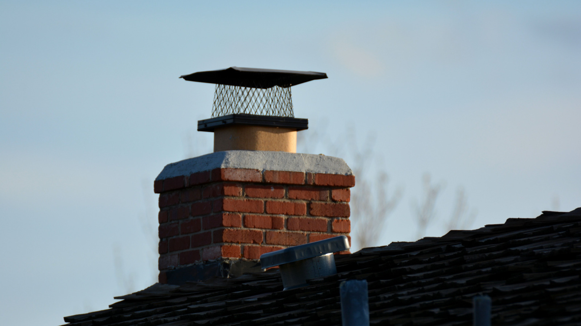 Brick chimney with black cap on a shingled roof against a pale blue sky
