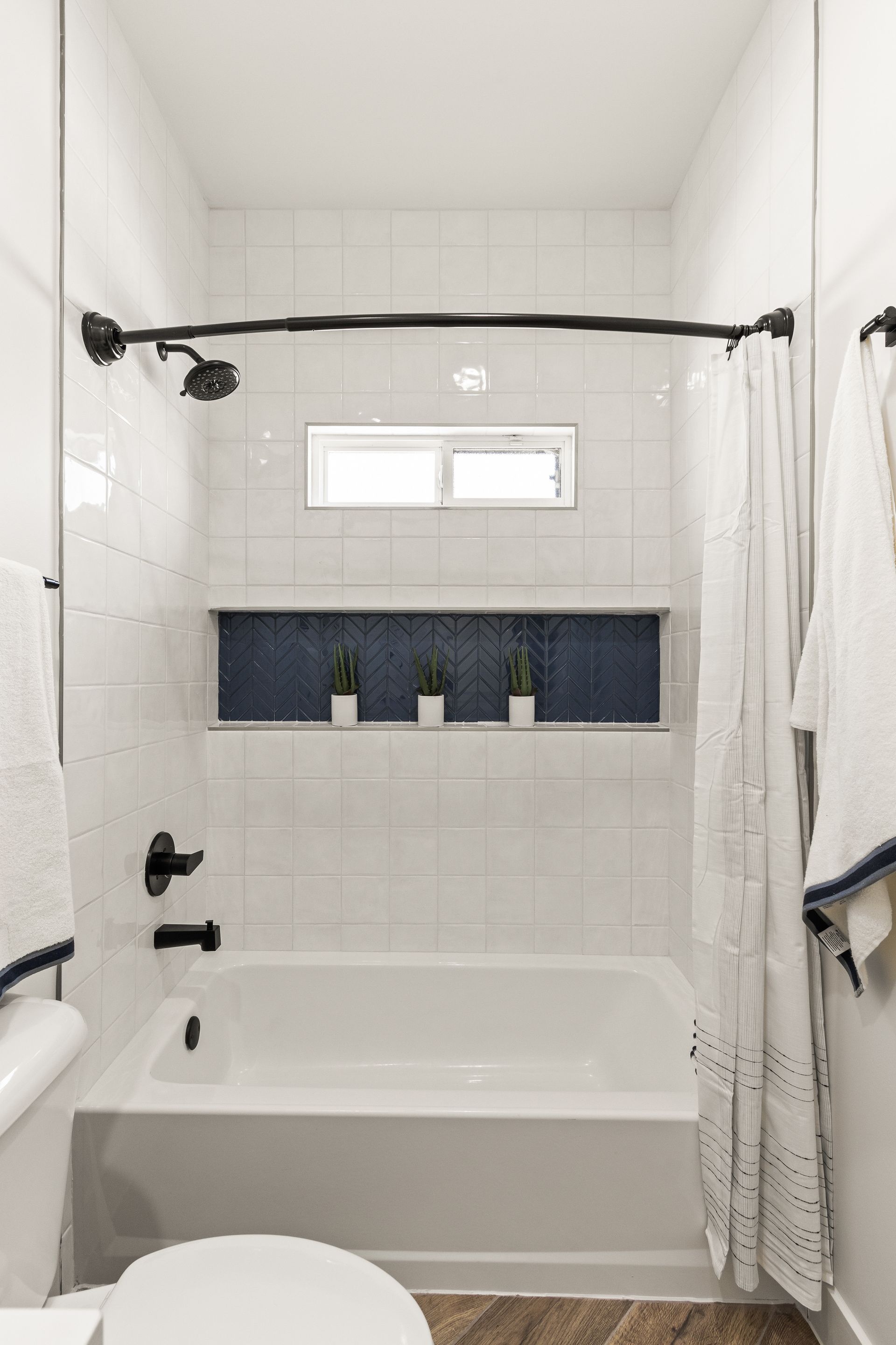 White tiled bathroom with a tub, showerhead, and blue inset with plants.