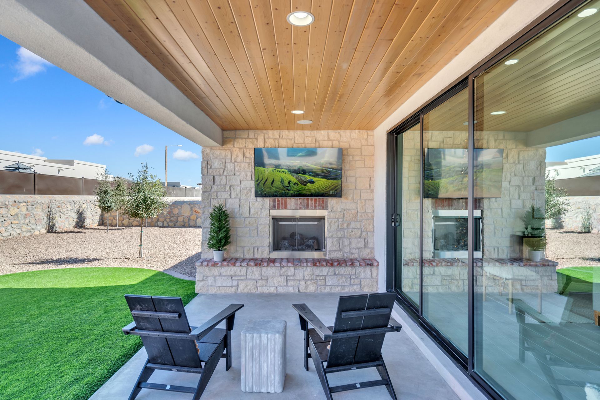 Outdoor patio with fireplace, TV, and seating under a wood-paneled ceiling, overlooking a grassy yard.