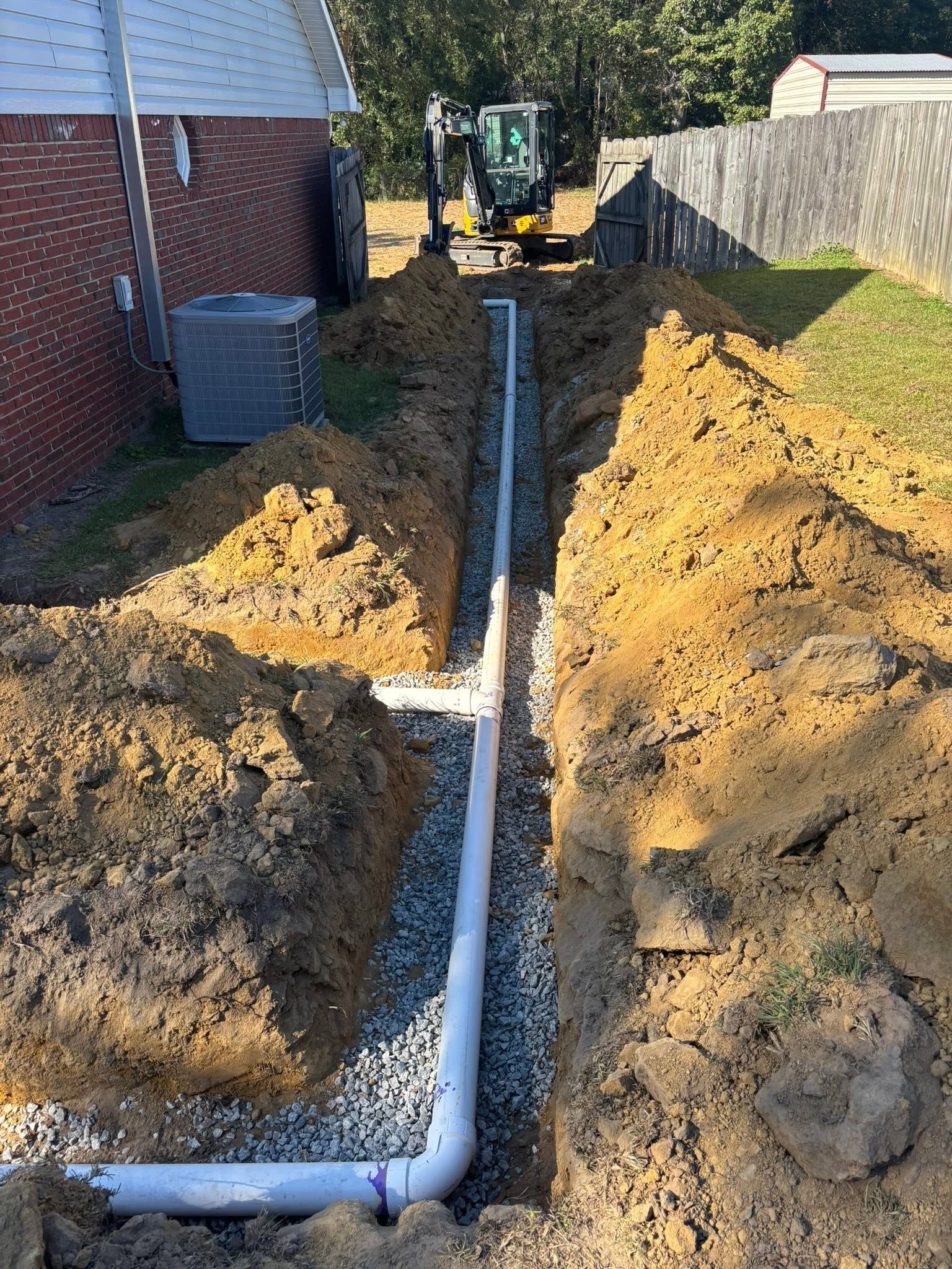 Trench with white PVC pipe being installed beside a house, with a backhoe in the background.