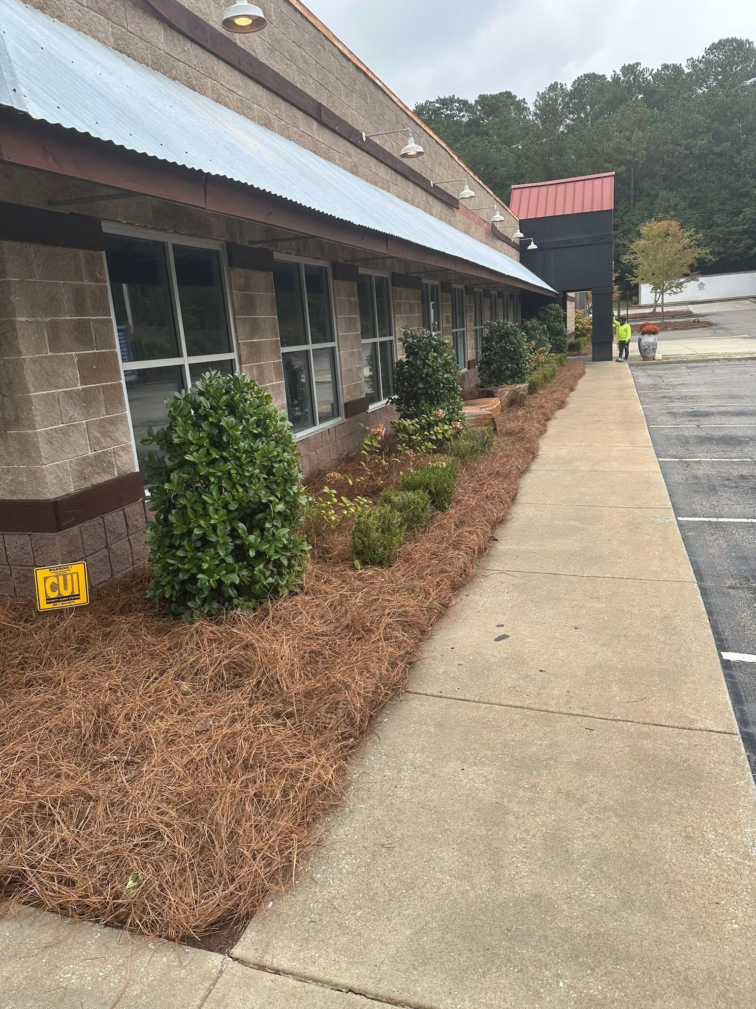 Sidewalk beside a brick storefront with shrubs, mulch, and a striped awning on a cloudy day