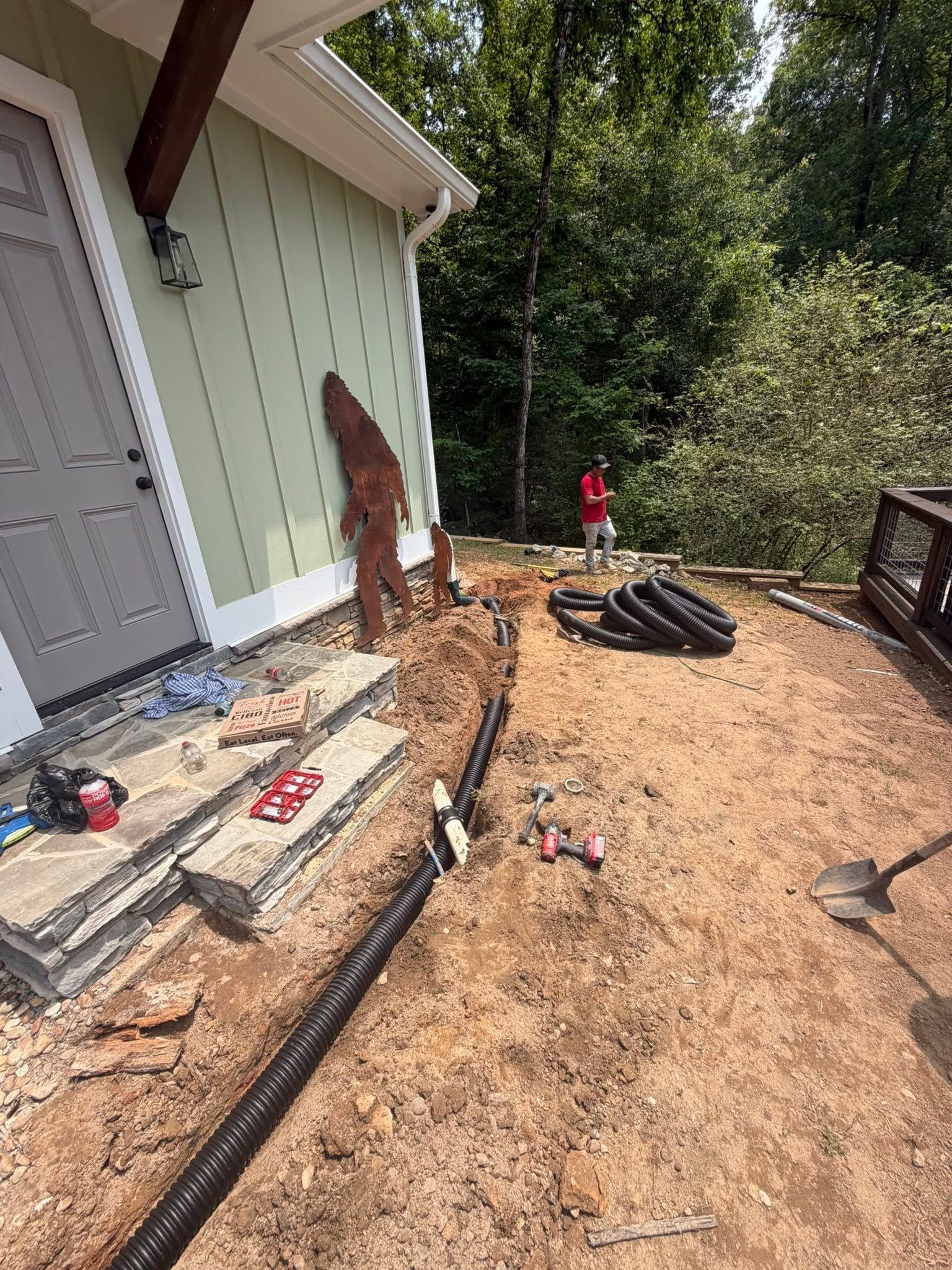 Backyard construction with dirt, pavers, black edging, and tools beside a house and trees