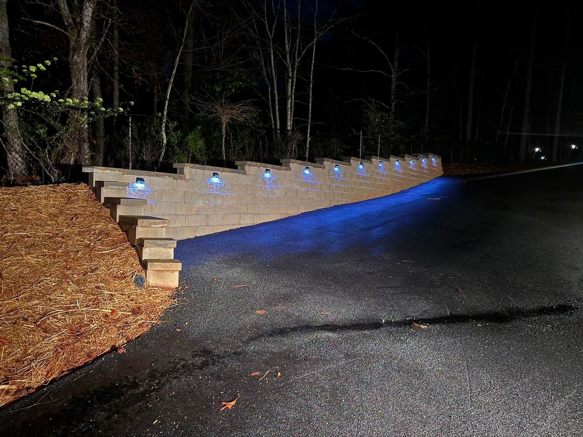 Dimly lit road at night beside a stone retaining wall with blue ground lights.