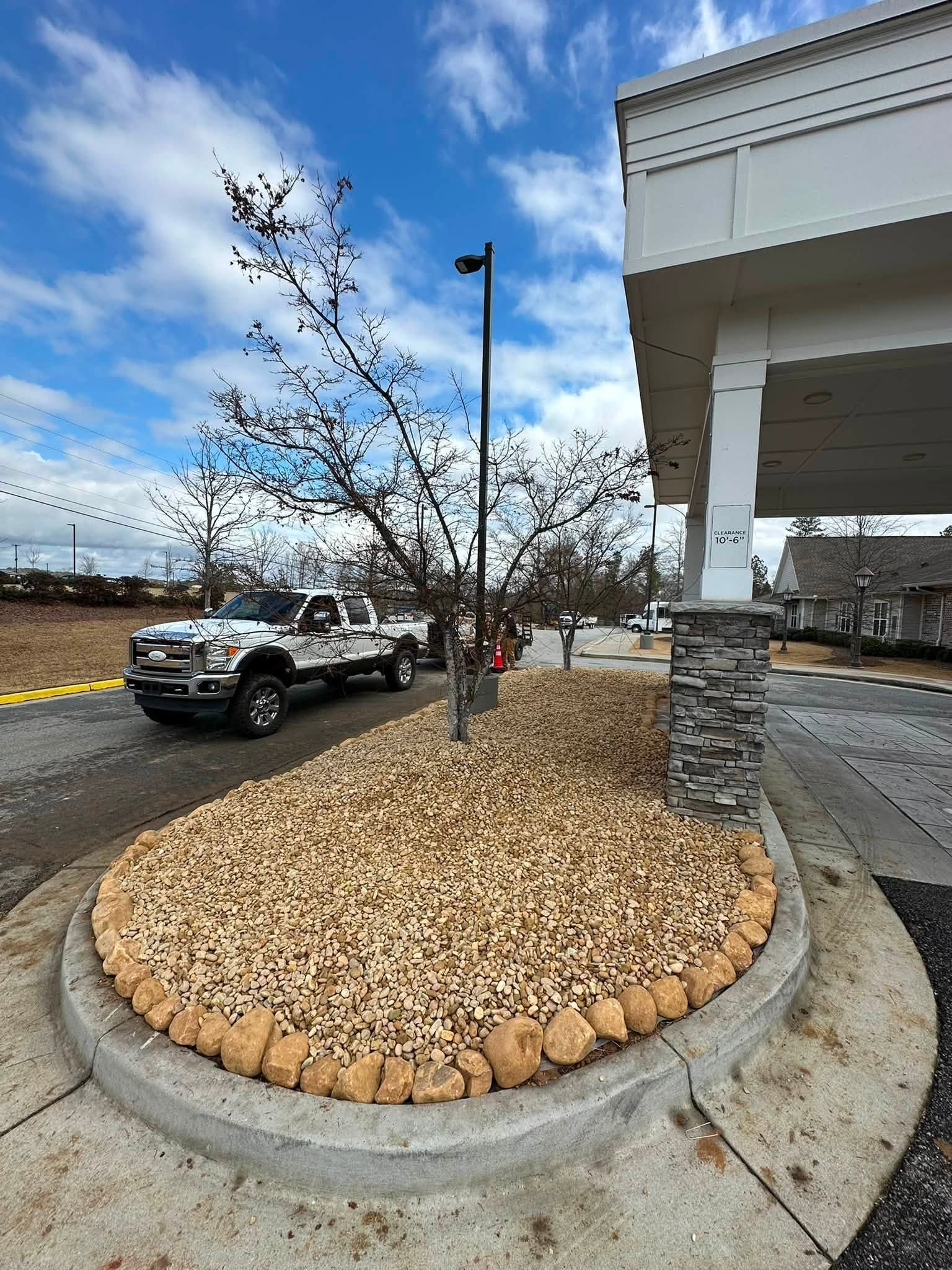 Empty landscaped curb island with leafless tree beside a building entrance and parked trucks under a blue sky