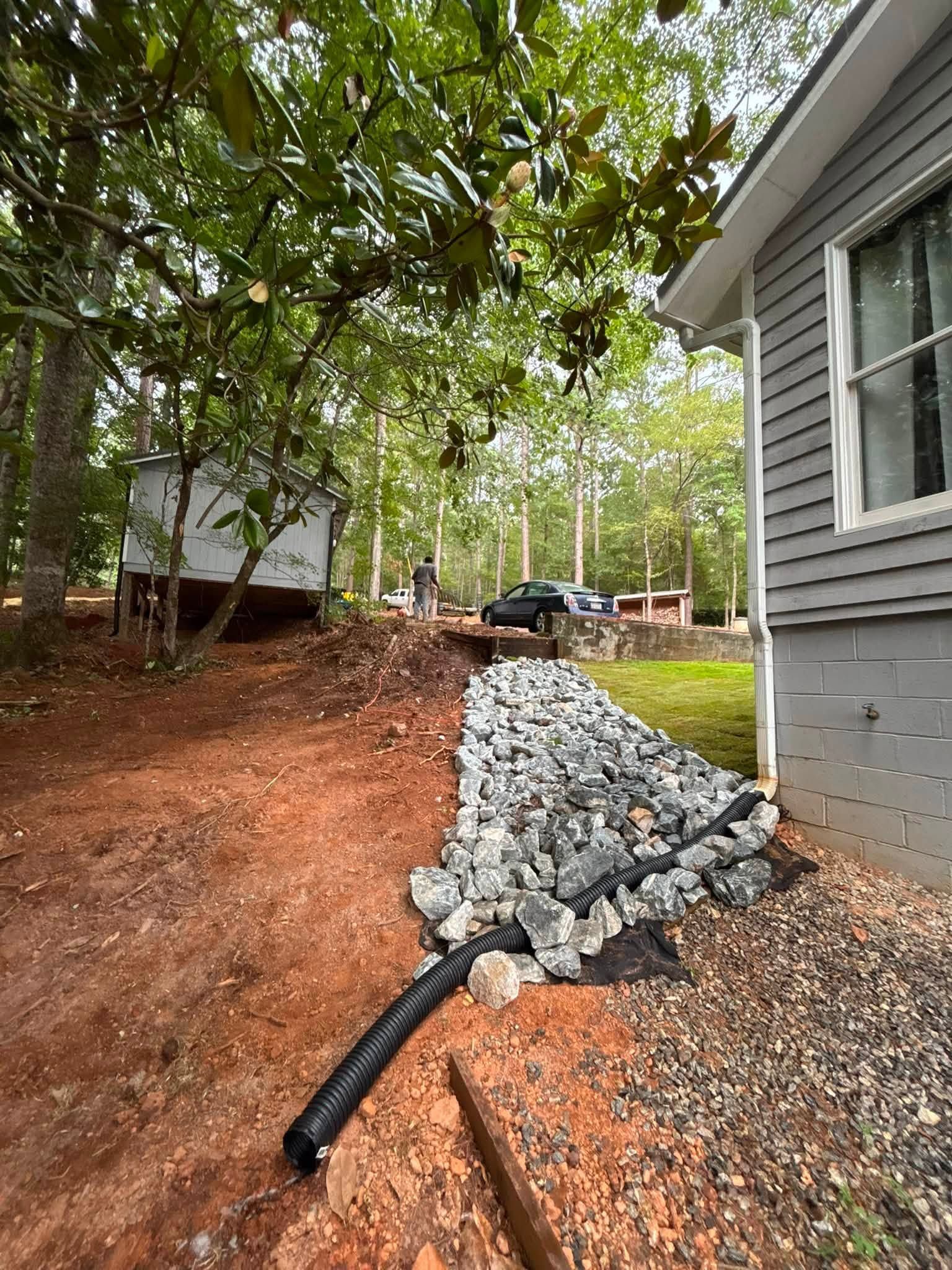 Black drainage pipe beside a house, lined with rocks, on a sloped wooded yard with red soil