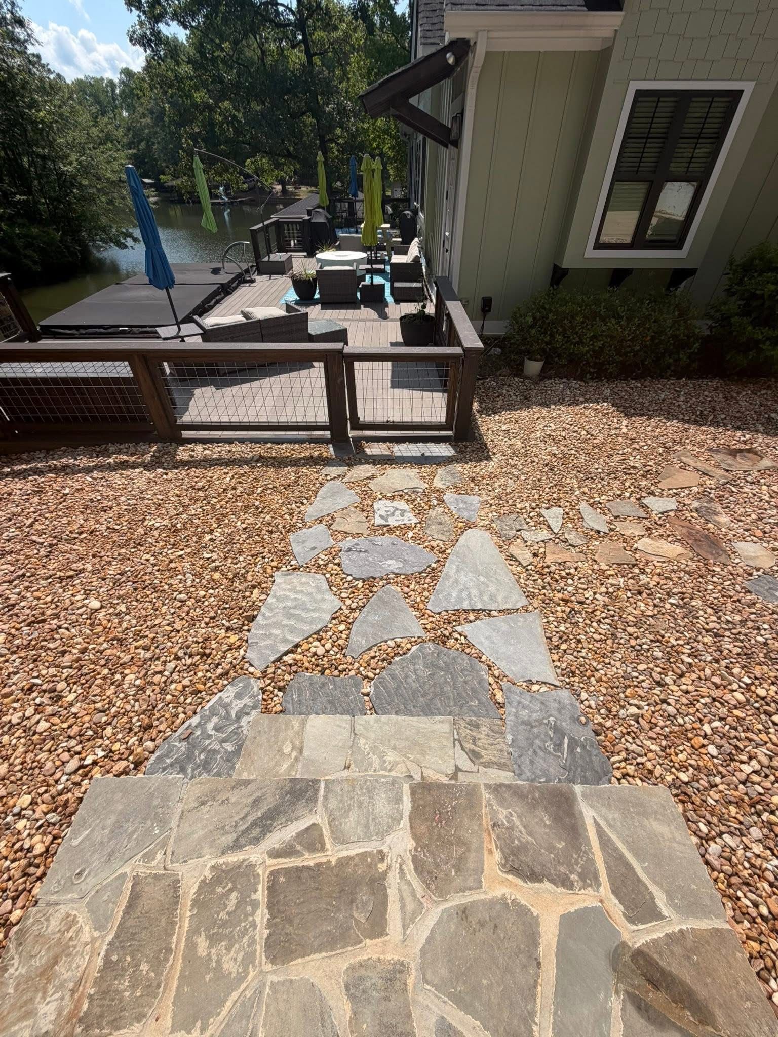 Stone walkway leading to a deck and house beside a lake, with gravel ground and trees.
