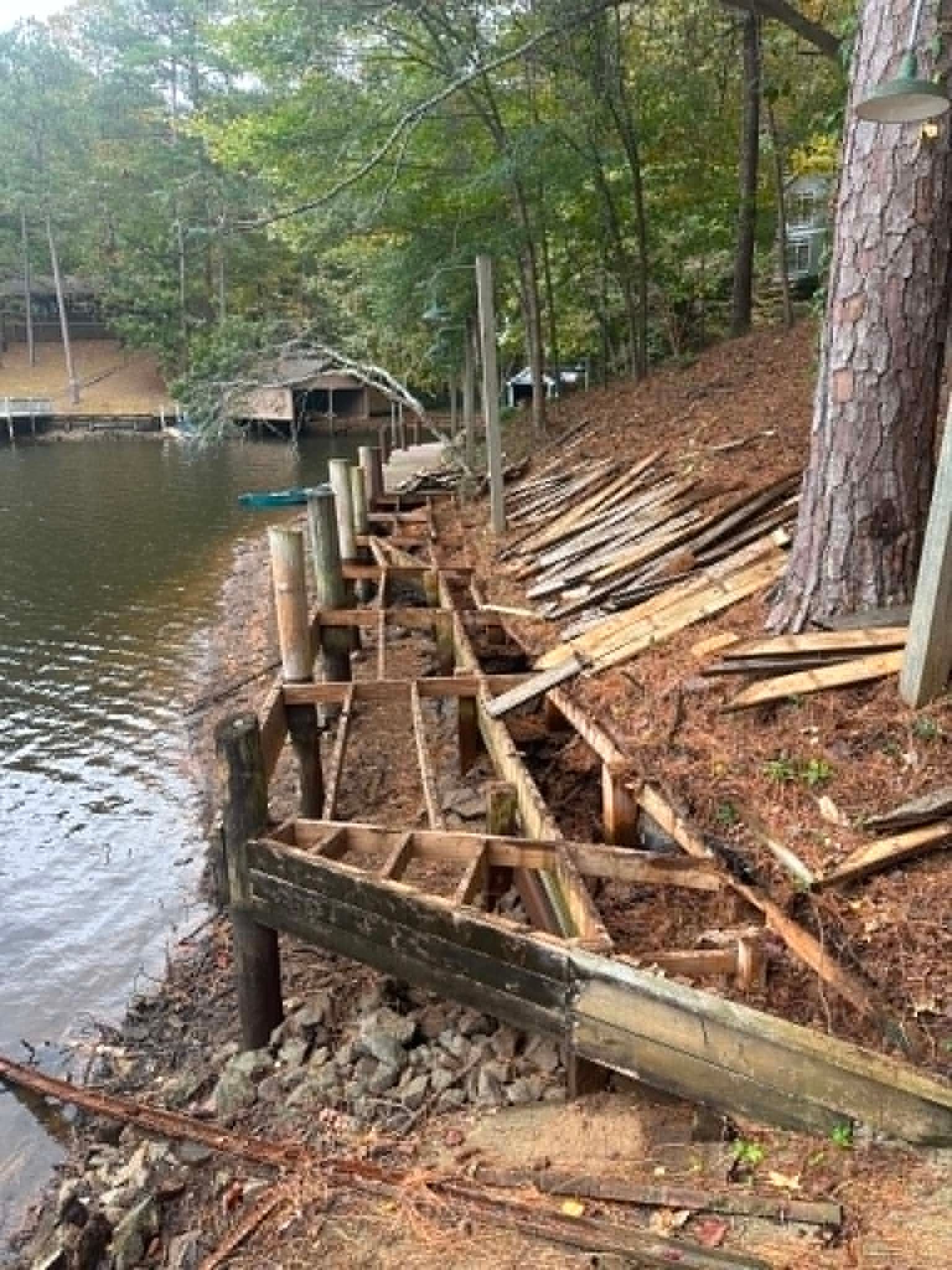 Collapsed wooden seawall along a wooded lakeshore, with exposed supports and eroded bank.