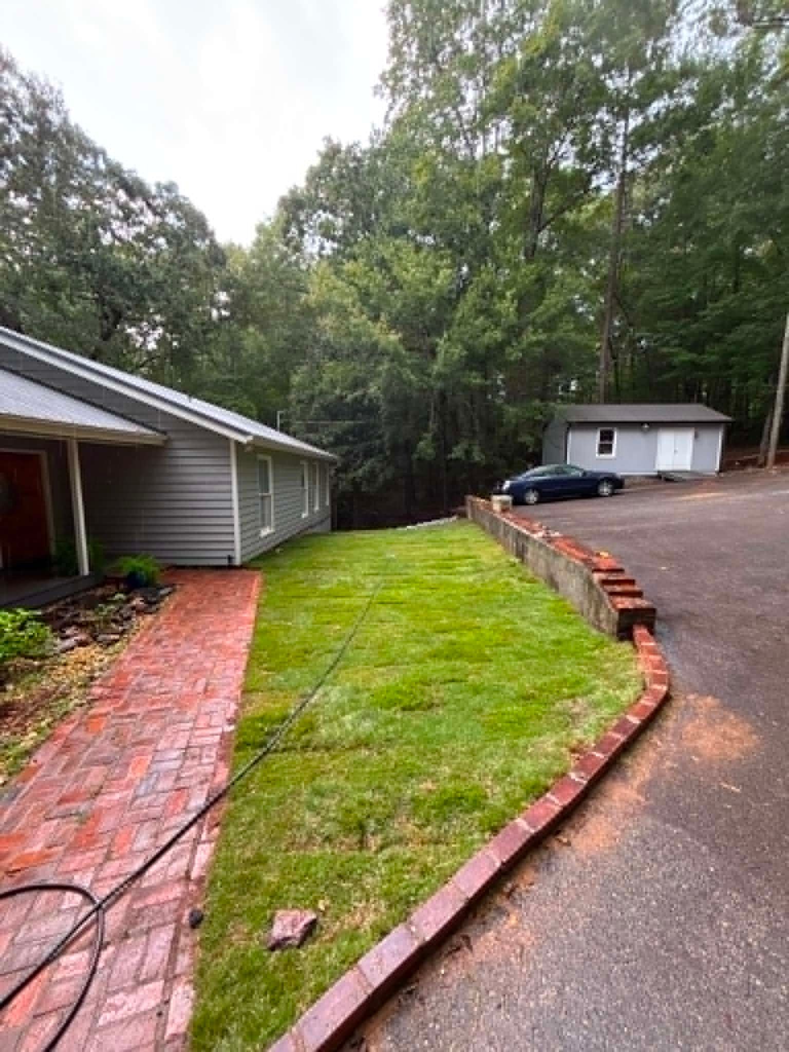 Brick walkway beside house leading to a driveway and small outbuilding, with a narrow grassy strip between.