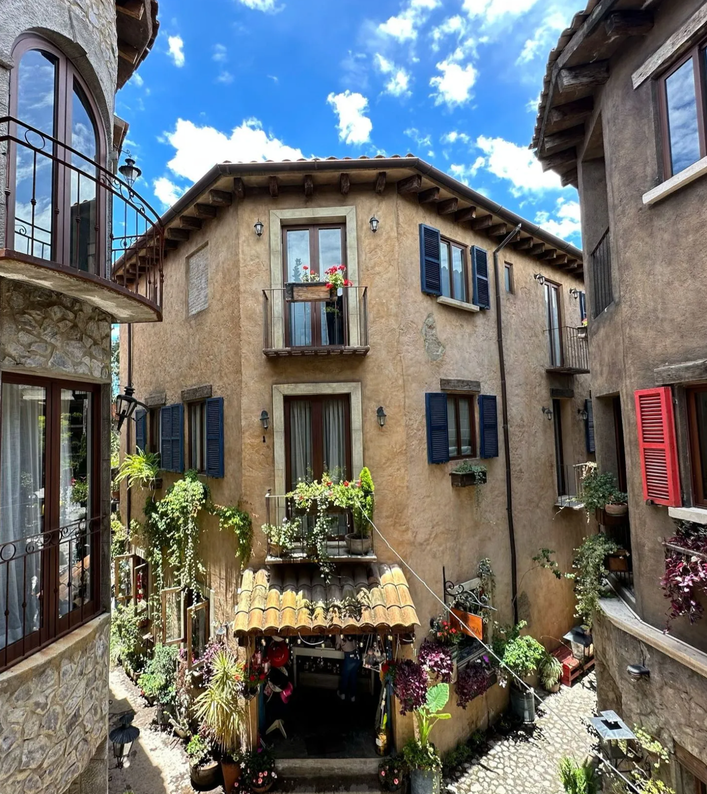 Narrow stone alley with rustic balconies, blue shutters, and potted flowers under a bright sky.