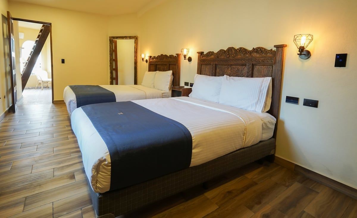 Bright hotel room with two queen beds, wood floors, wall lamps, and a doorway to a balcony.