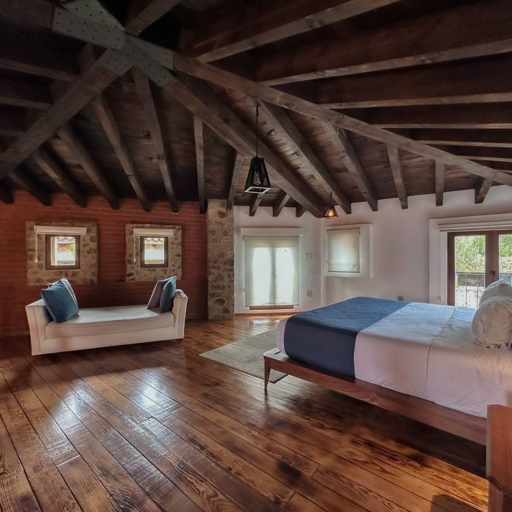 Rustic attic bedroom with exposed wood beams, brick wall, bed, sofa, and sunlight through windows