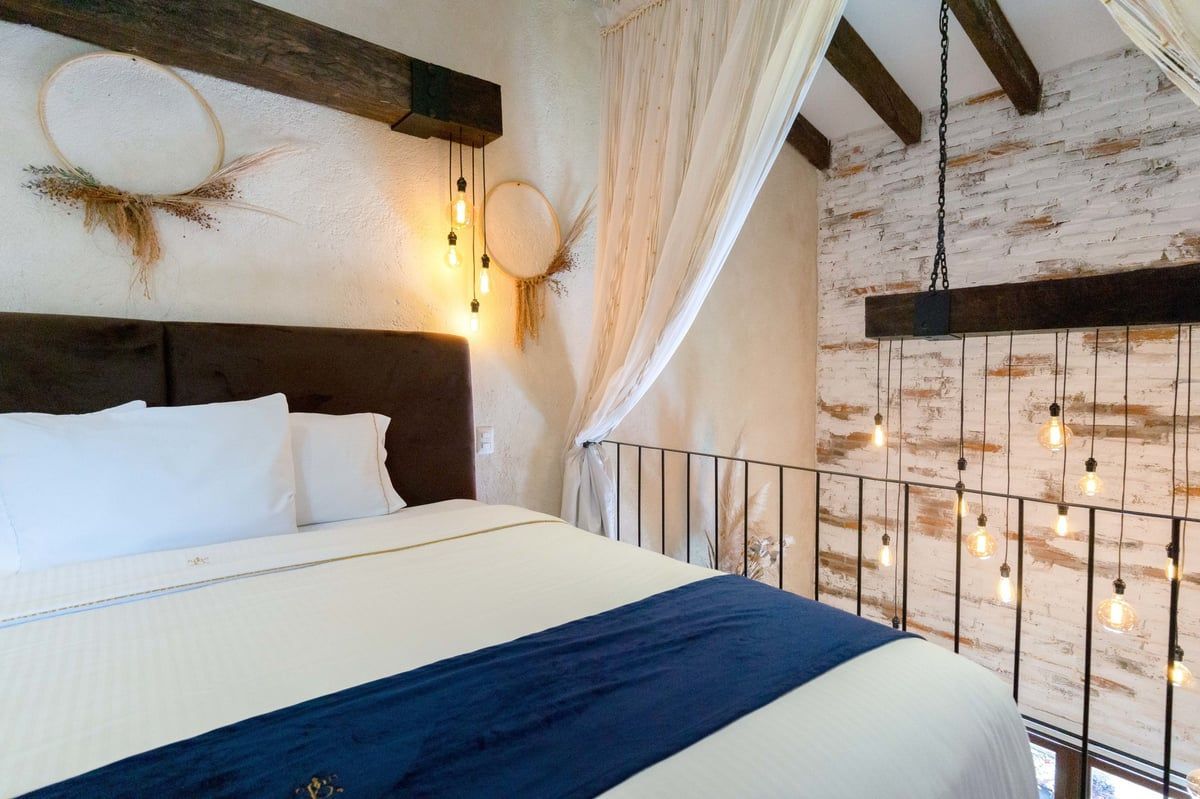Cozy bedroom with white bedding, dark blue throw, brick walls, and sheer canopy drapes near a window