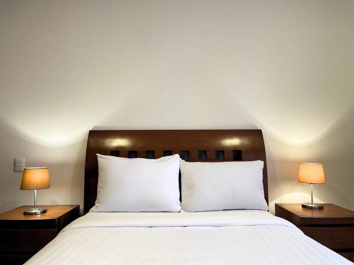 A neatly made bed with white linens, two bedside lamps, and a softly lit bedroom wall.
