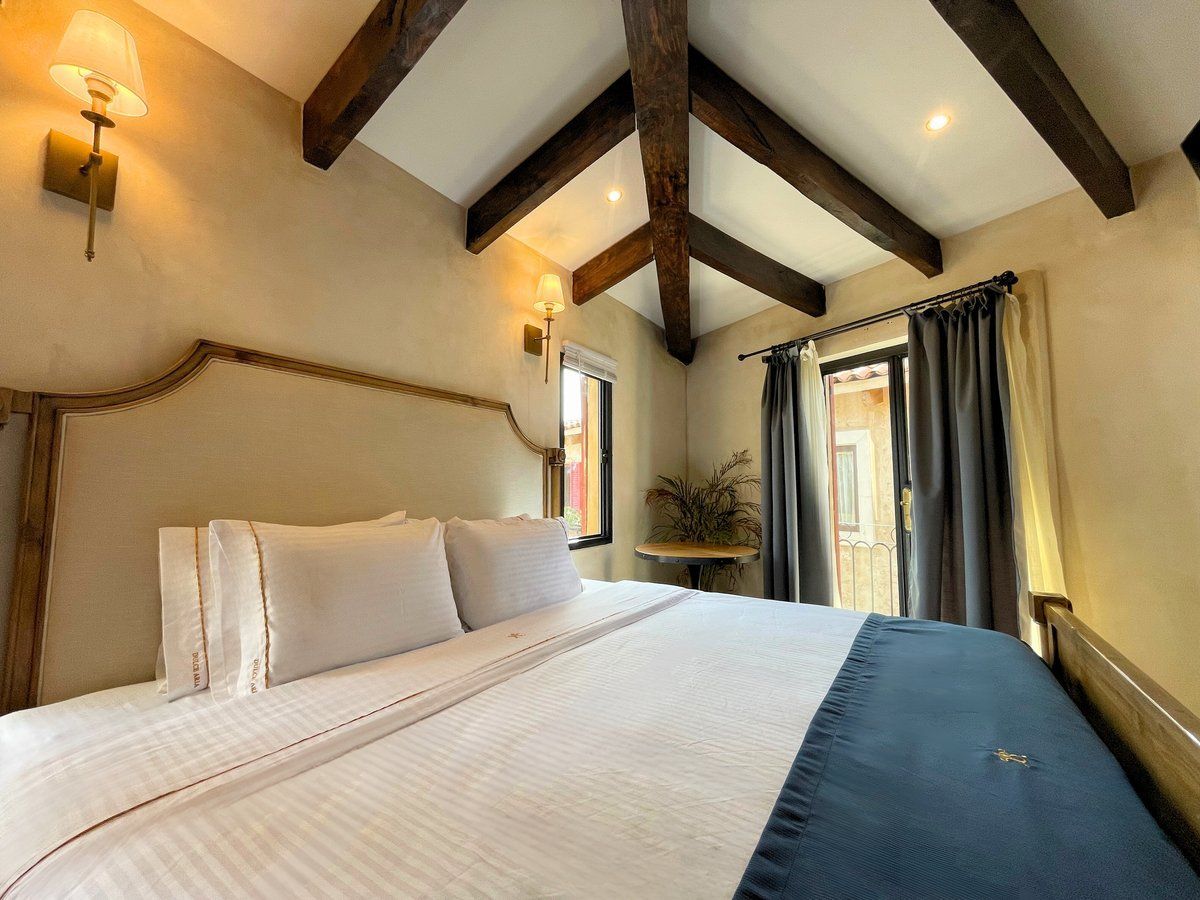 Cozy bedroom with exposed wood beams, a large bed, wall sconces, and a window with curtains