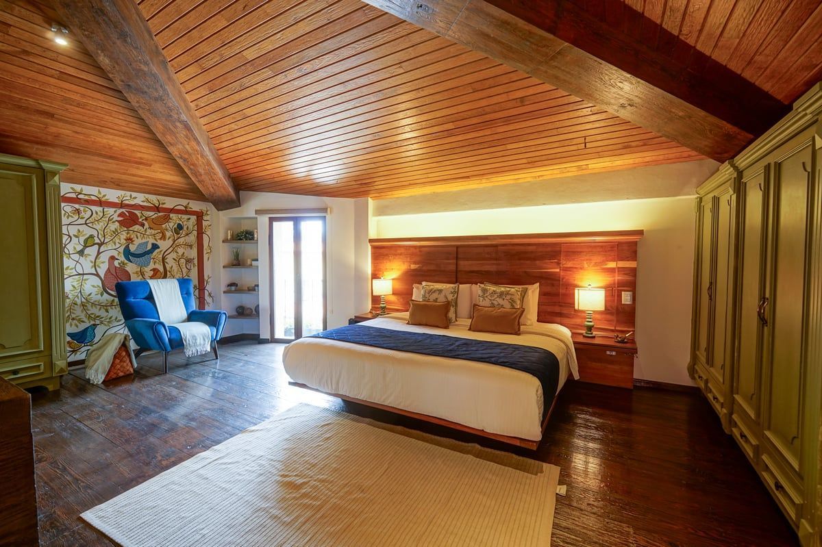 Rustic bedroom with carved wooden ceiling, large bed, and blue armchair beside a window.