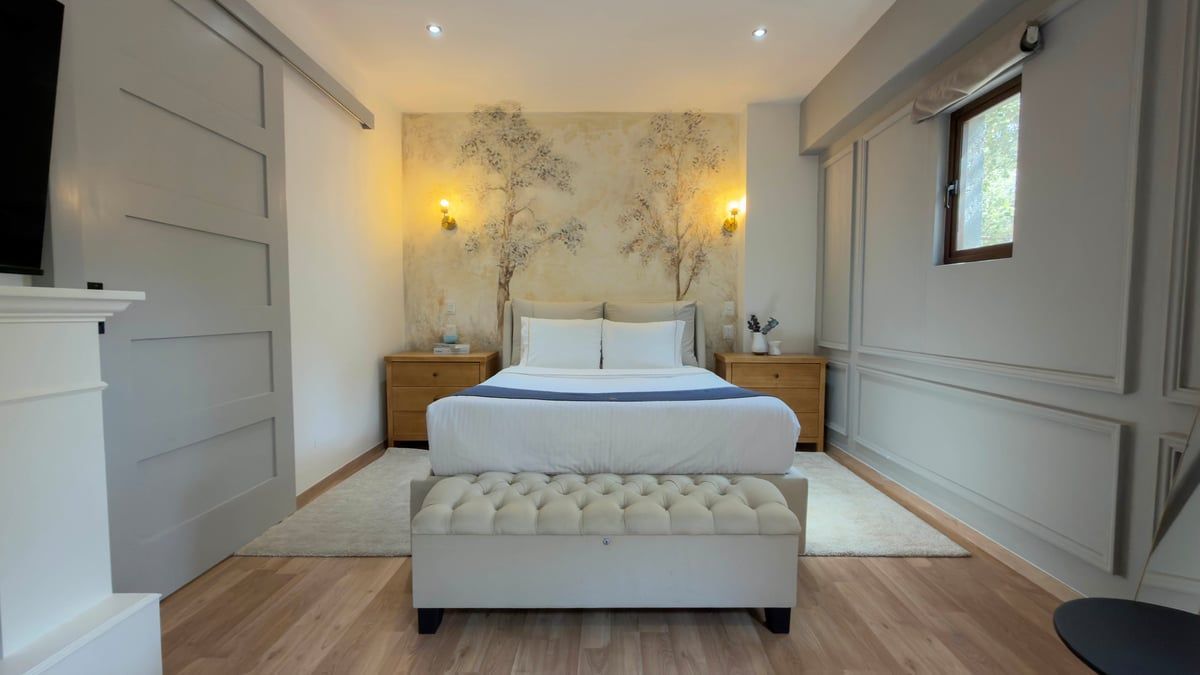 Bright bedroom with a neatly made white bed, tufted bench, wall mural, and soft lighting.