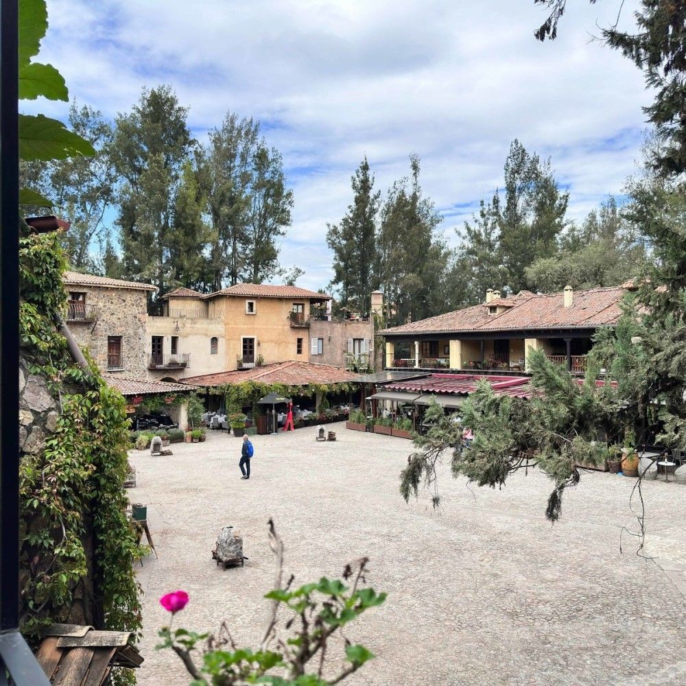 Courtyard with rustic buildings, trees, and a few people under a cloudy sky