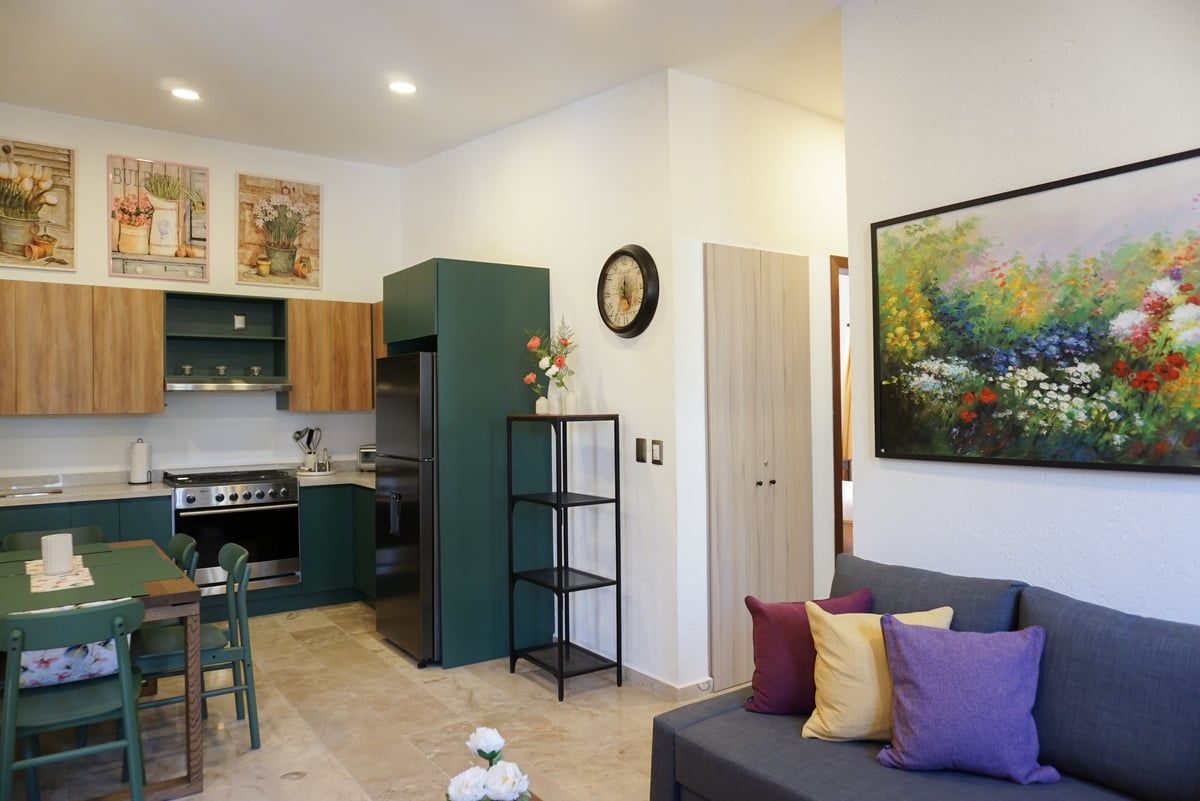 Modern open-plan living room and kitchen with green cabinets, gray sofa, colorful wall art, and potted plant.