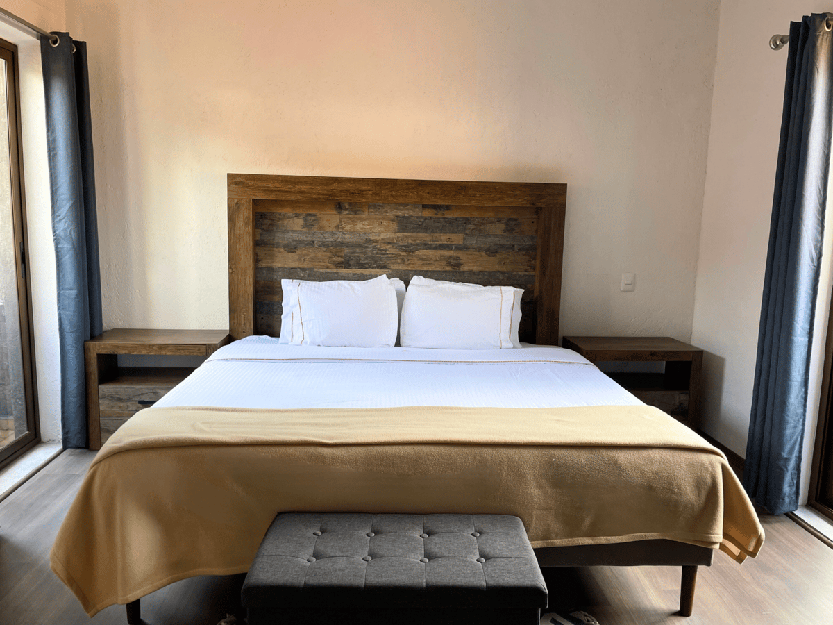 Bedroom with a king bed, wooden headboard, bench, and tall windows with dark curtains