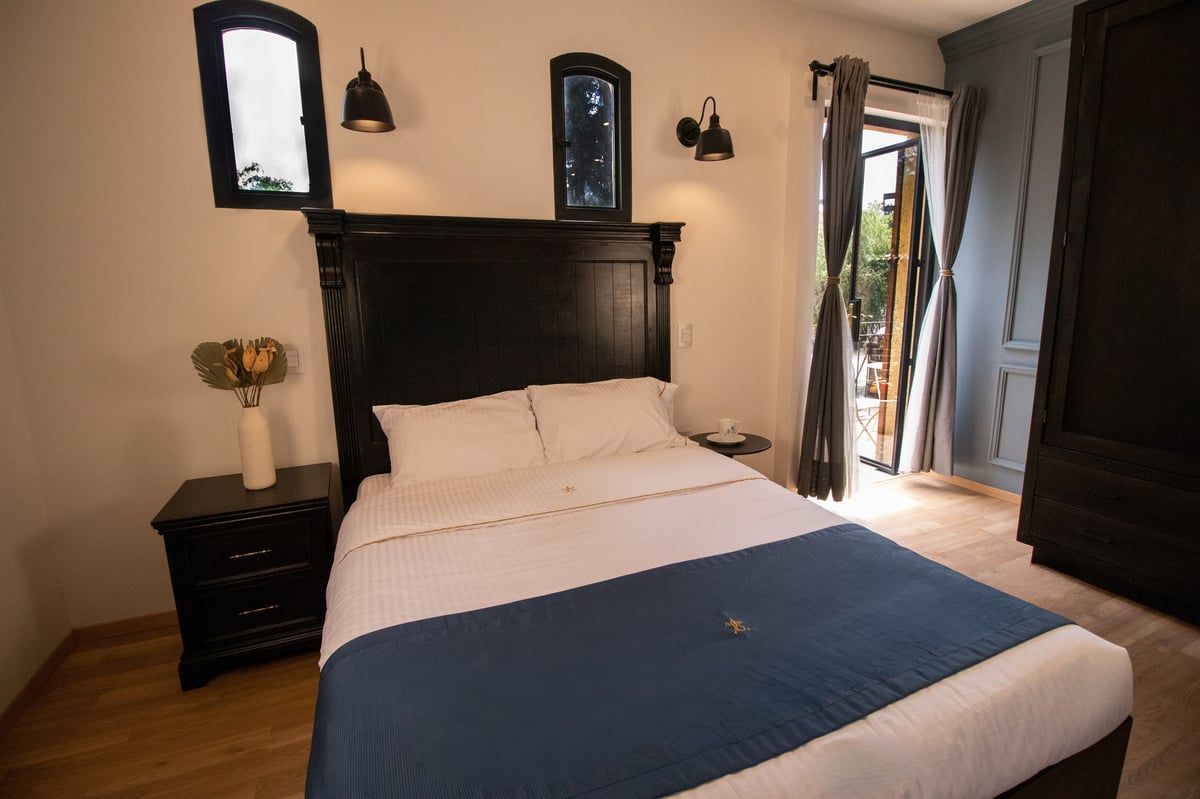 Bedroom with a dark wood bed, blue runner, bedside table, wall sconces, and an open door to a balcony.