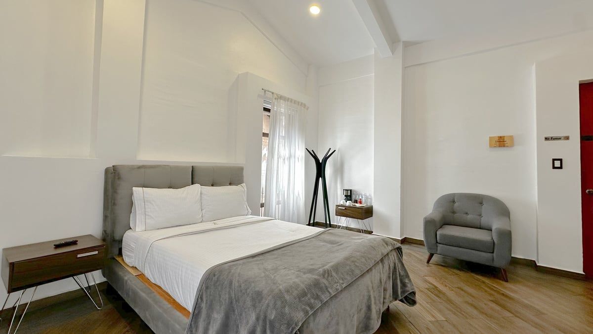 Bright bedroom with a made bed, gray chair, wood floor, and white walls.