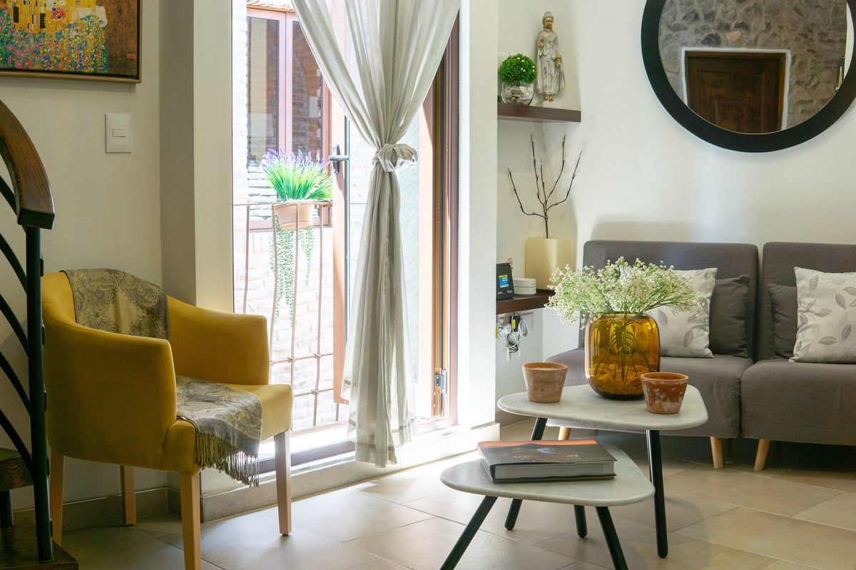 Bright living room with mustard armchair, gray sofa, round mirror, and coffee table by a sunlit balcony door
