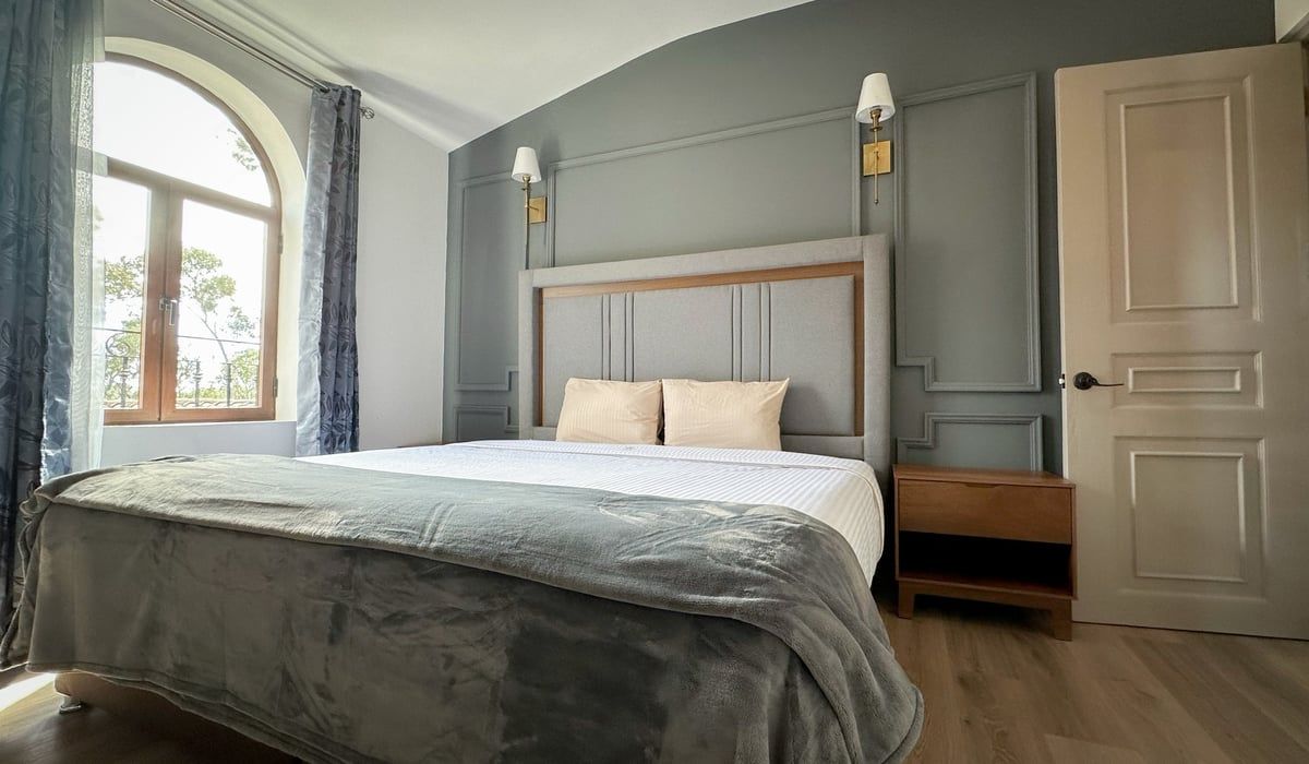 Bright bedroom with a large bed, arched window, blue-gray walls, and beige door.