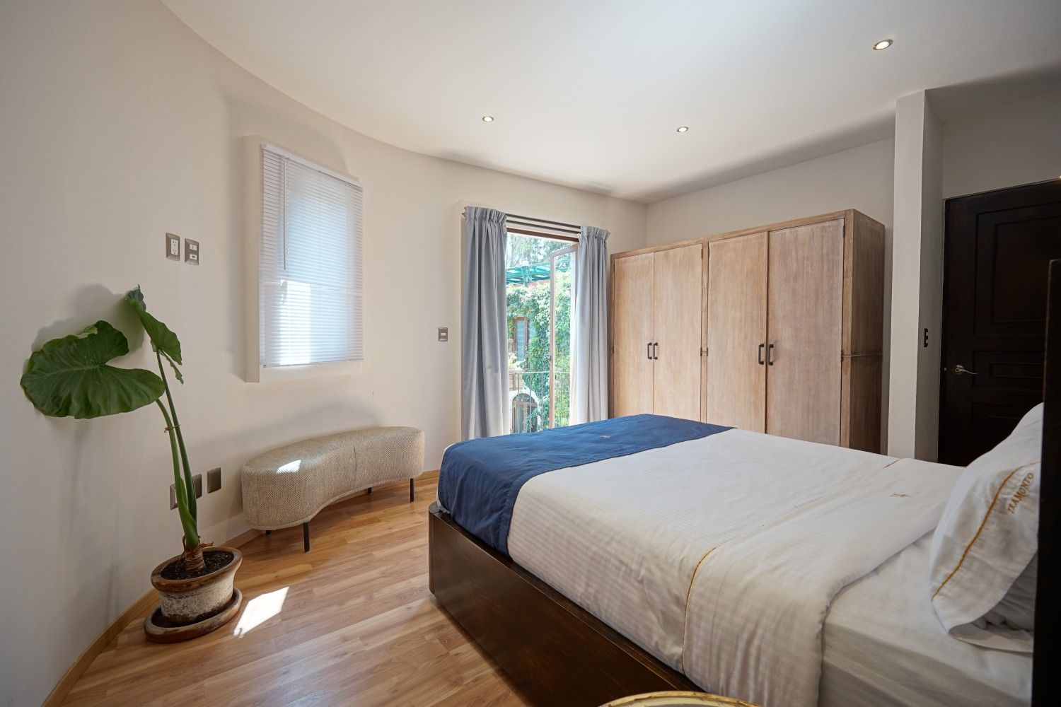 Bright bedroom with a bed, wardrobe, plant, bench, and open door to a sunlit balcony.