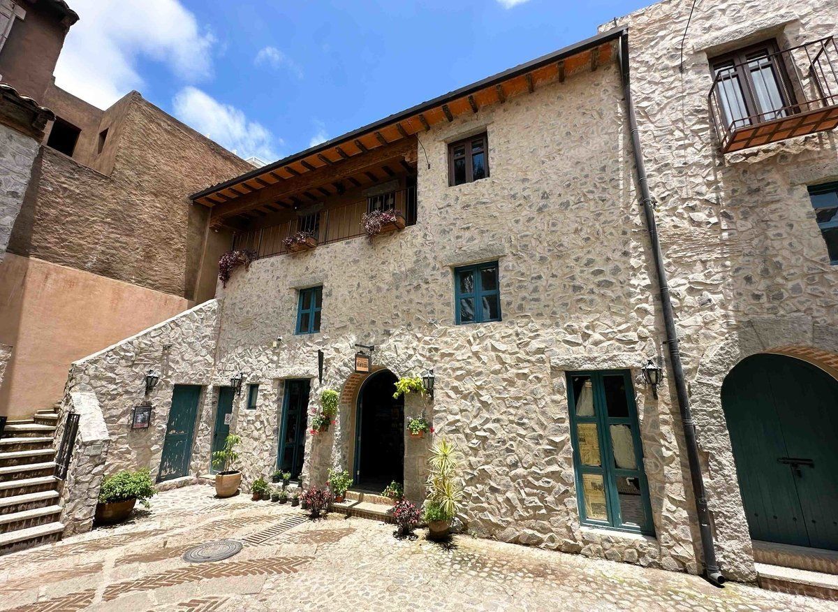 Stone courtyard with rustic old buildings, green shutters, and an arched doorway under a blue sky