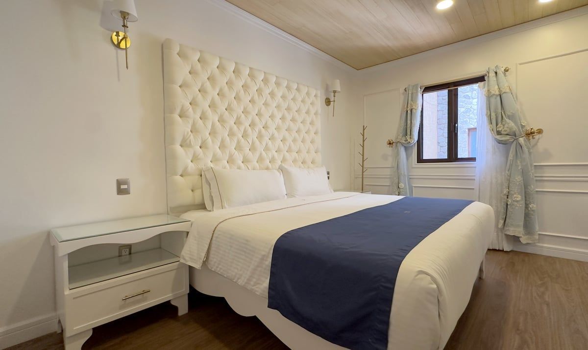 Bright bedroom with white bed, blue runner, tufted headboard, and a window with sheer curtains.