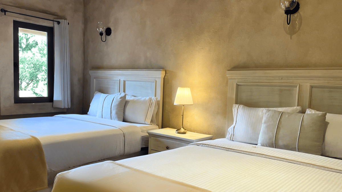 Two twin beds in a beige bedroom with a lit lamp and a window