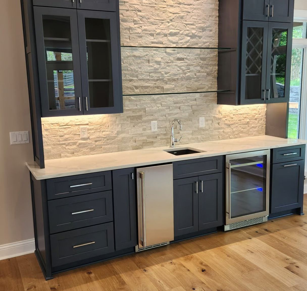Modern kitchen bar with dark cabinets, stone backsplash, white countertop, sink, mini fridge, and wine cooler.