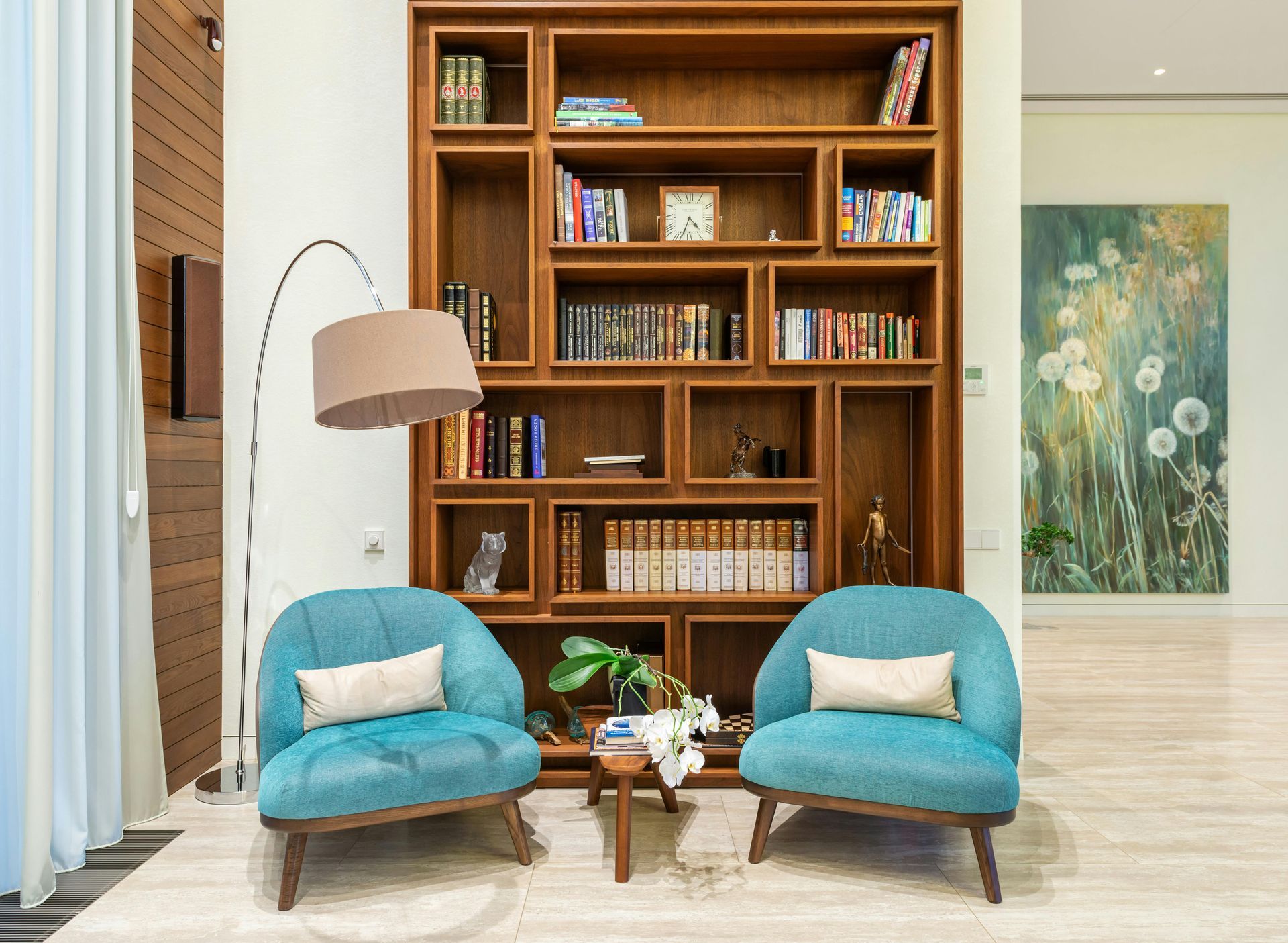 Modern living room with blue chairs, bookshelves, floor lamp, and floral wall art