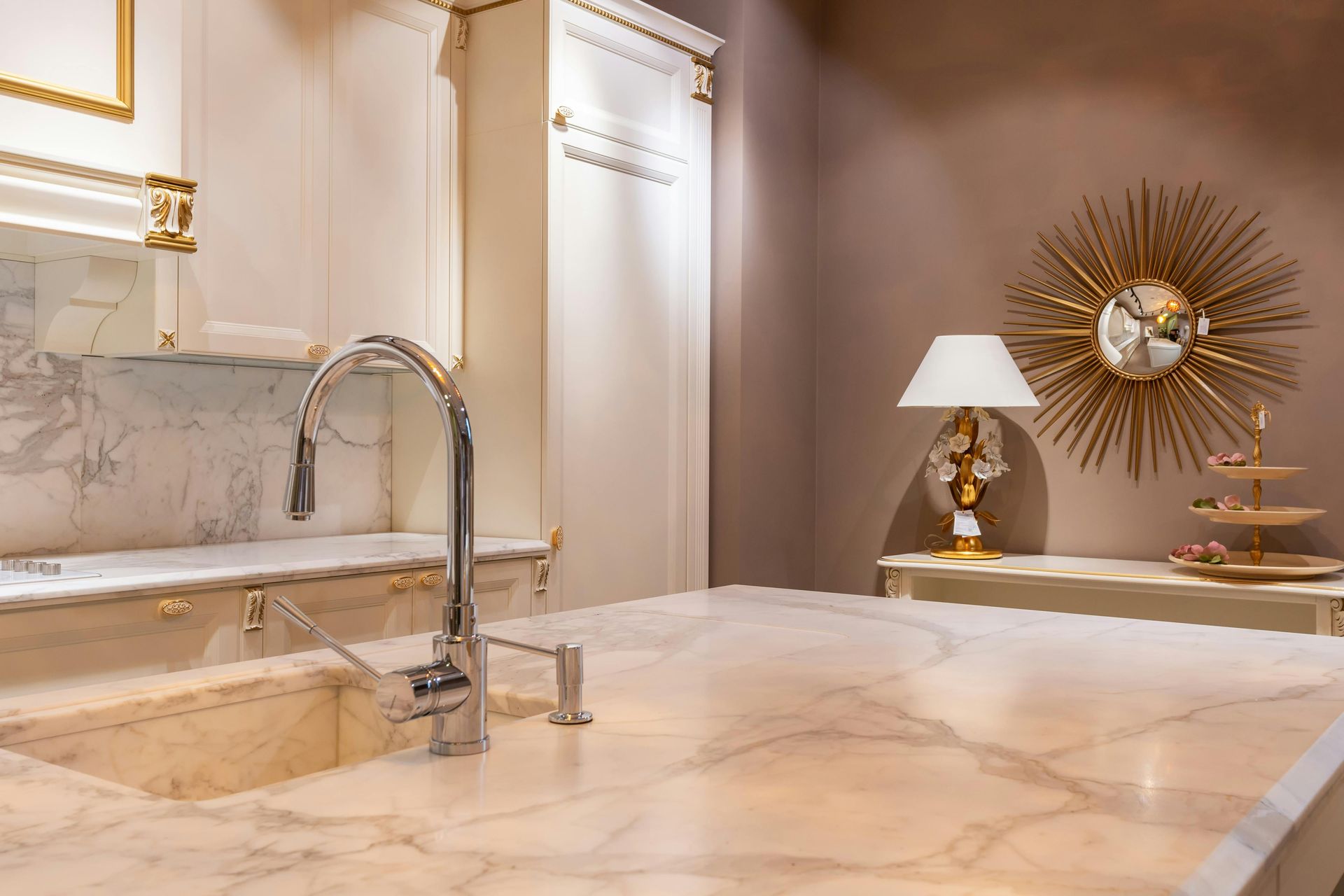 Elegant marble bathroom with a modern faucet, gold accents, and a table lamp beside a sunburst wall mirror