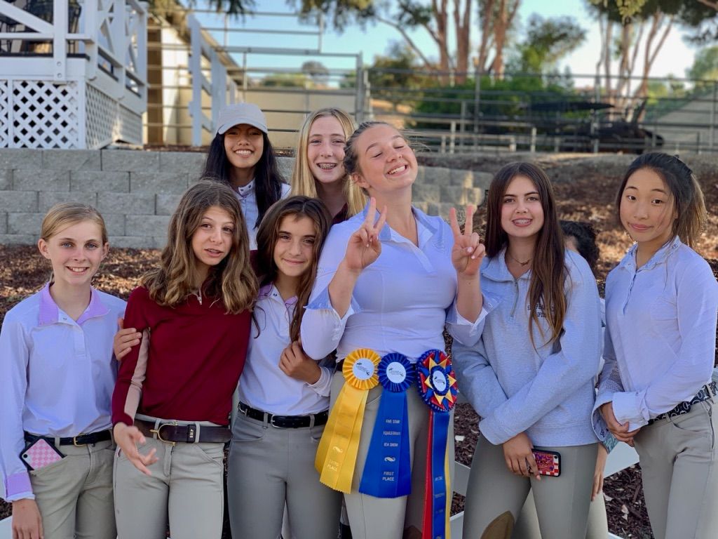 Group of six smiling people outdoors, standing by a blue ribbon trophy at what looks like a sports celebration