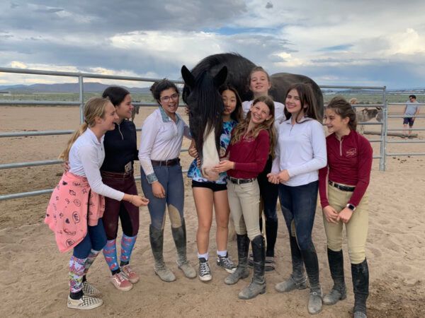 Group of people standing with a black horse on a sandy beach, smiling and posing together.