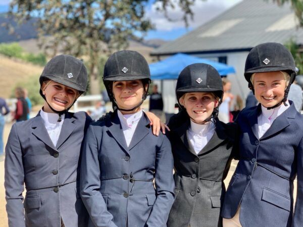 Four people in riding helmets and formal jackets stand together outdoors, smiling at the camera.