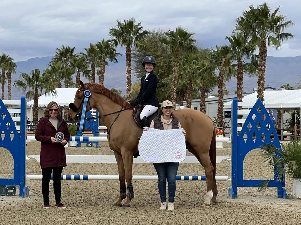 Horse show winner on tan horse with blue jumps, rider and two people holding award ribbon outdoors